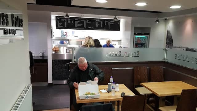A man sits at a wooden table enjoying fish and chips in a modern restaurant with a menu board and staff behind the counter.