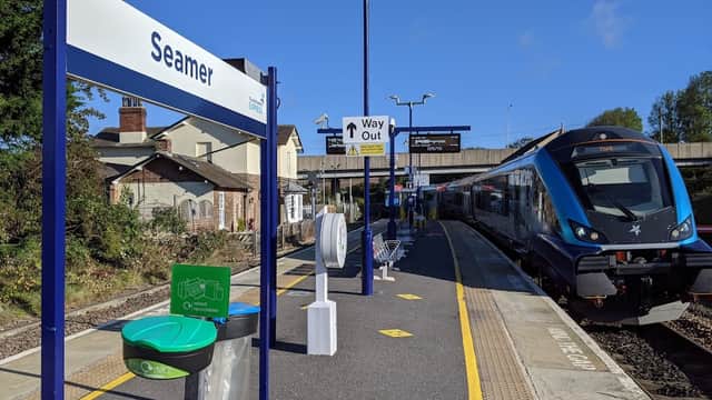 Seamer train station platform with signage, a train, and a green hand sanitizer station under a clear blue sky.