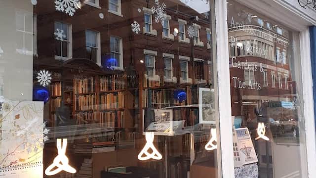 Bookshop window displaying books, snowflake decorations, and warm lighting, with reflections of nearby buildings.