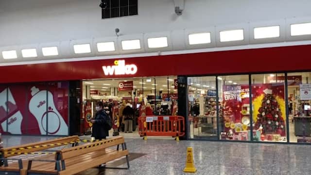 Entrance to Wilko store with festive decorations, a bench, and caution sign on the floor in a shopping center.