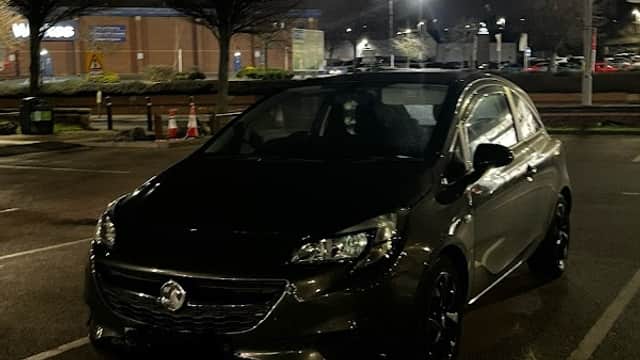 A black car parked in a lot at night, with streetlights and trees in the background.