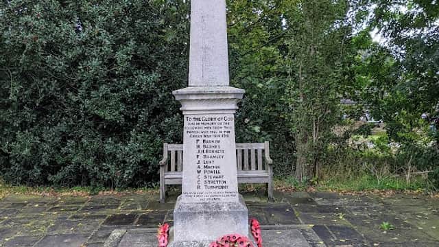 War memorial with an obelisk, surrounded by greenery and benches, adorned with poppy wreaths and a plaque.