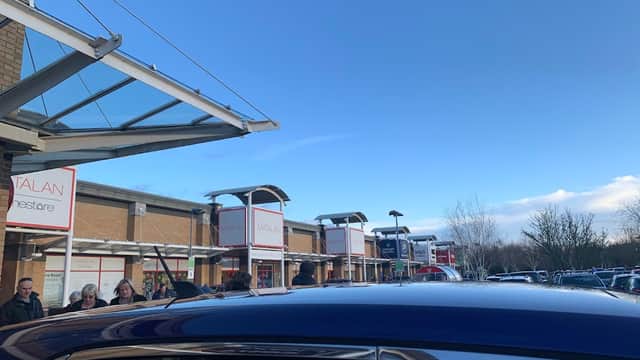 Shops and car park at St James Retail Park under a clear blue sky, with people walking and parked cars visible.