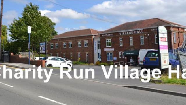 Village hall with red brick facade, sign reading "Charity Run Village Hall," parked cars, and green trees nearby.