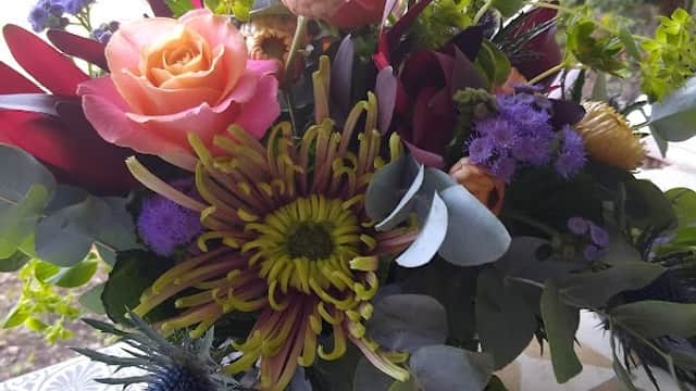 Vibrant floral arrangement with pink roses, yellow chrysanthemums, and purple accents in a decorative container.
