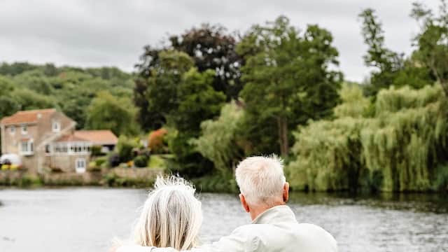 A couple sits on a wooden bench by a tranquil lake, surrounded by lush greenery and a cloudy sky.