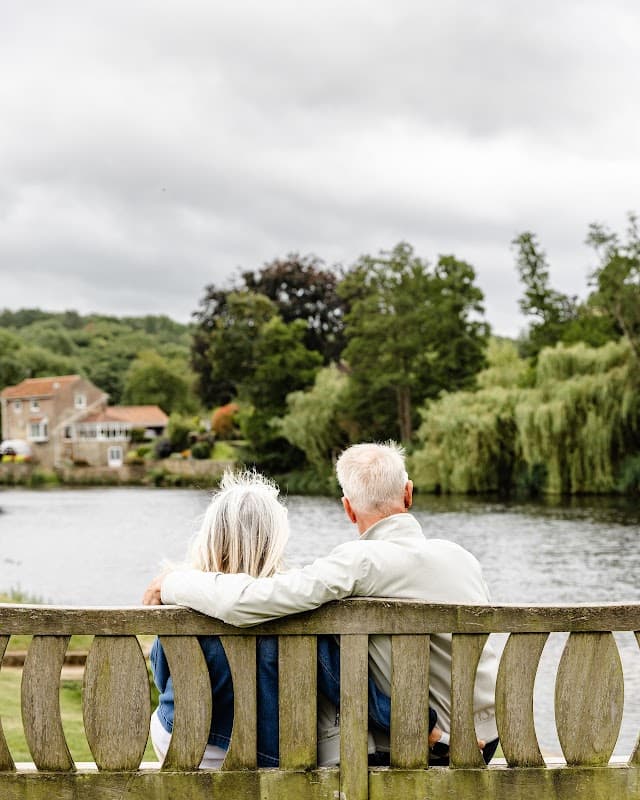 A couple sits on a wooden bench by a tranquil lake, surrounded by lush greenery and a cloudy sky.