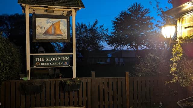 The Sloop Inn sign features a painting of a sailboat, surrounded by trees and a cozy beer garden at dusk.
