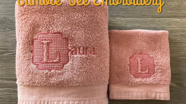Pink towels with embroidered initials "L" and "Laura" displayed on a wooden surface, with "Bumble Bee Embroidery" text above.