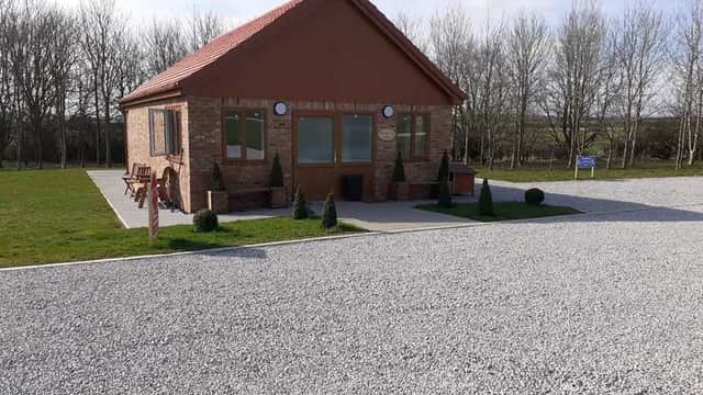 Quaint brick building with a red roof, surrounded by gravel and small shrubs, set against a backdrop of trees.
