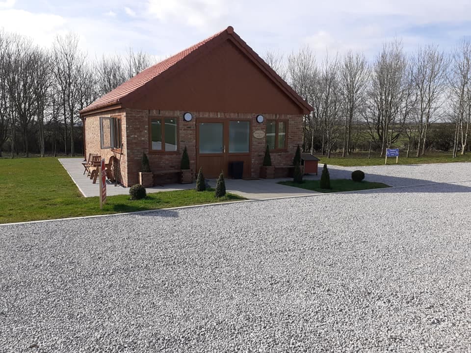 Quaint brick building with a red roof, surrounded by gravel and small shrubs, set against a backdrop of trees.