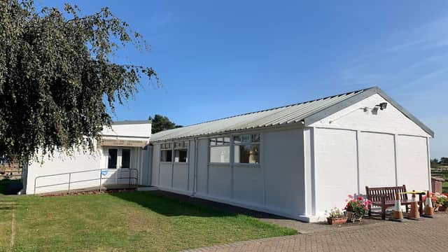White village hall with a sloped roof, surrounded by green grass and a bench, under a clear blue sky.