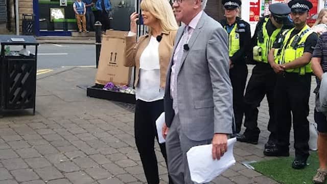 A man in a suit and a woman carrying a bag walk through Birstall Market Place, with police officers in the background.