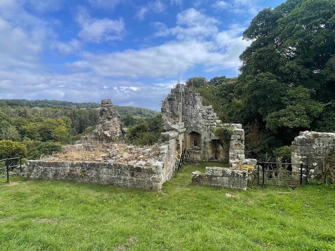 Ruined stone structure surrounded by lush greenery and trees under a partly cloudy sky in Mulgrave Woods.