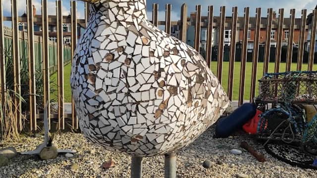 Mosaic seagull sculpture stands on a pebbled area, with a grassy field and wooden fence in the background.