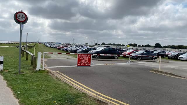 Park and Ride area with parked cars, a no parking sign, and wind turbines in the background under a cloudy sky.