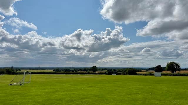 Green sports field with a goalpost, under a blue sky filled with clouds, overlooking a rural landscape.