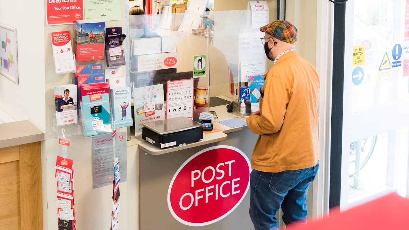 Customer at a counter in New Earswick Post Office, surrounded by brochures and a prominent red "POST OFFICE" sign.