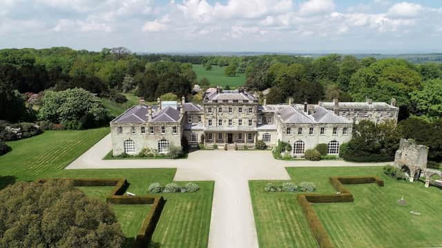 Elegant stone mansion surrounded by manicured lawns and lush greenery under a bright blue sky.
