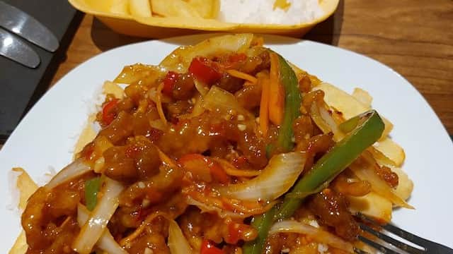A plate of stir-fried chicken with vegetables and fries, next to a takeout container with rice and more fries.