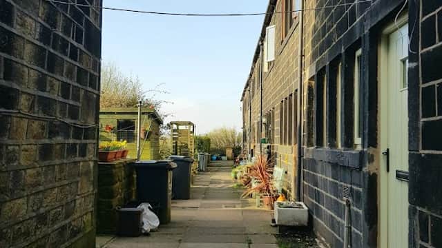 Narrow pathway between stone buildings, with planters and bins, leading to a garden area under a clear blue sky.