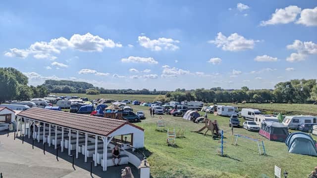 Busy campsite with tents and caravans under a bright blue sky, surrounded by green fields and trees.