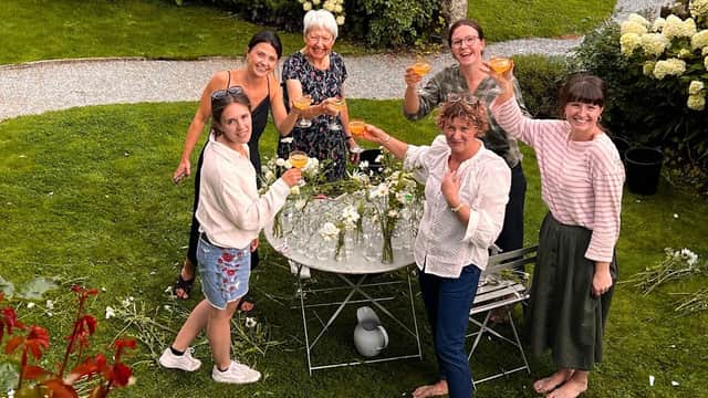 A group of six women celebrating with drinks in a garden, surrounded by flowers and greenery.