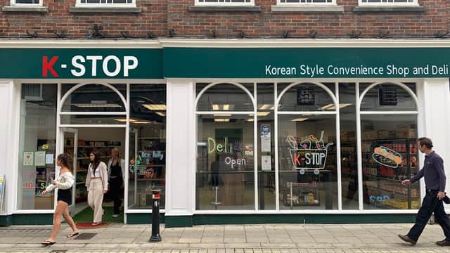 K-STOP store front with large windows, displaying colorful products and a "Deli Open" sign, people walking by.