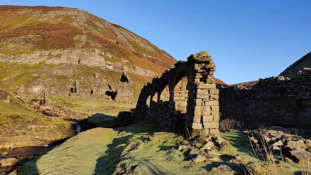 Stone ruins with archways beside a stream, set against a backdrop of green hills and clear blue sky.