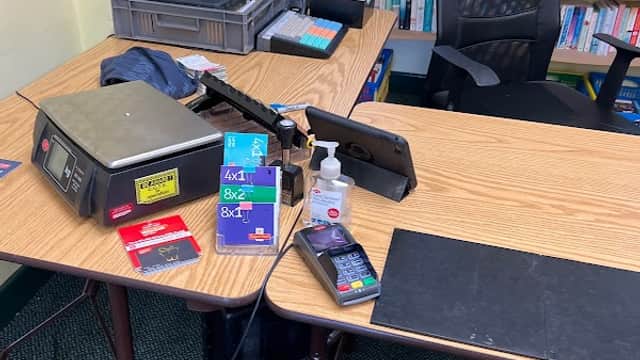 A small post office counter with a cash register, payment terminal, hand sanitizer, and a chair in front of a bookshelf.