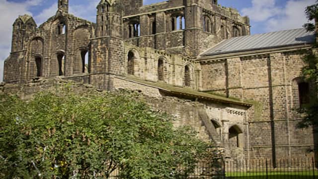Medieval stone ruins with arched windows and towers, surrounded by lush green grass and trees under a blue sky.
