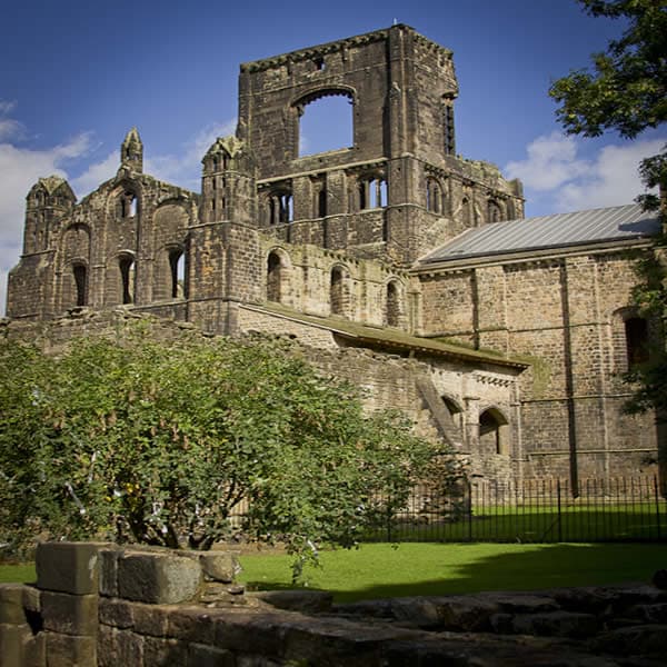 Medieval stone ruins with arched windows and towers, surrounded by lush green grass and trees under a blue sky.