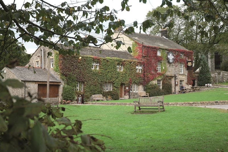 The Lister Arms pub features ivy-covered stone walls, wooden benches, and a lush green lawn in Kirkby Malham, Yorkshire.