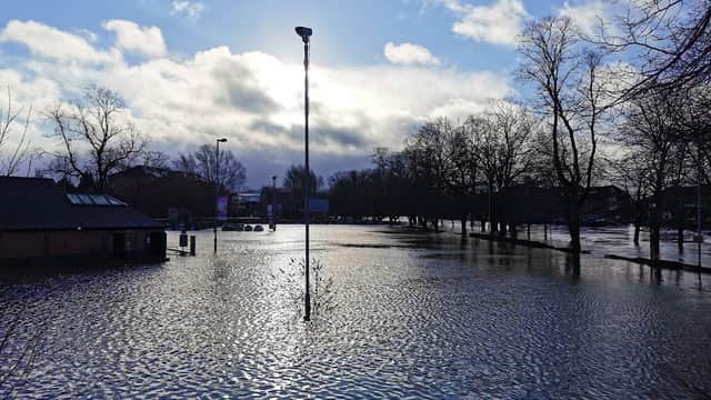 Flooded car park with standing water, trees in the background, and a cloudy sky with sunlight breaking through.