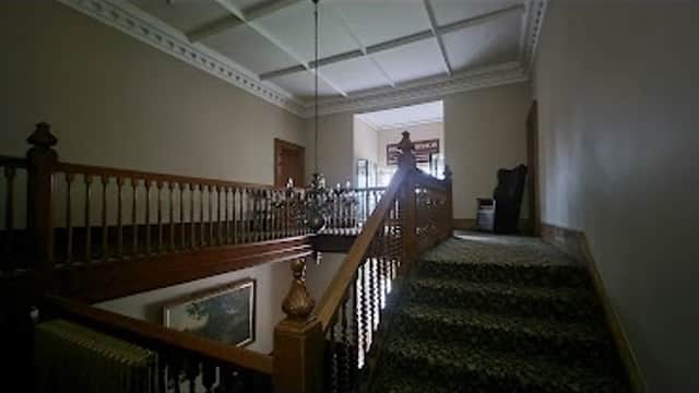 Staircase with wooden railing and carpet, leading to a well-lit room with large windows in a historic building.