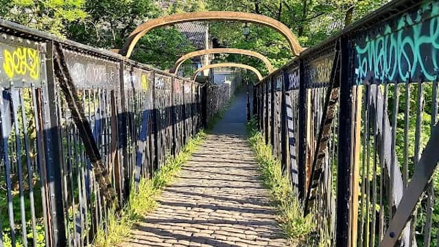 Footpath leading through a bridge surrounded by greenery, with graffiti on the railings and sunlight filtering through trees.