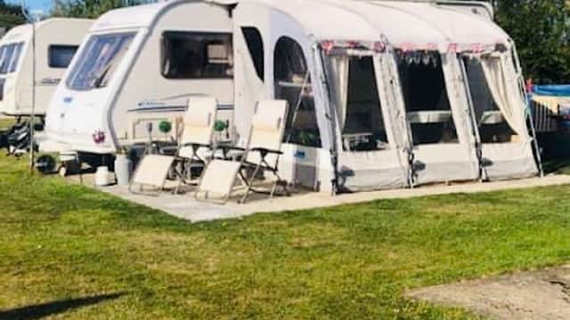 Caravan with an awning and outdoor chairs on a grassy area, surrounded by trees under a clear blue sky.