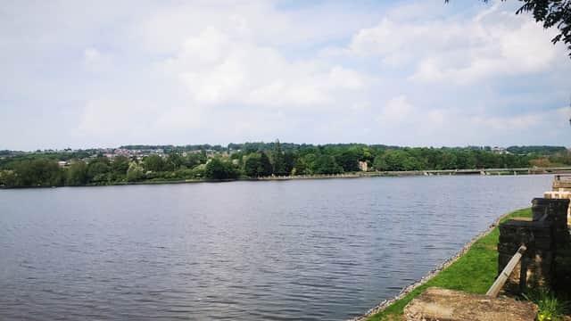 Scenic view of a lake surrounded by greenery under a cloudy sky, with a distant bridge and buildings visible.