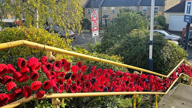 Colorful knitted poppies adorn a yellow railing along a staircase, with cars and houses in the background.