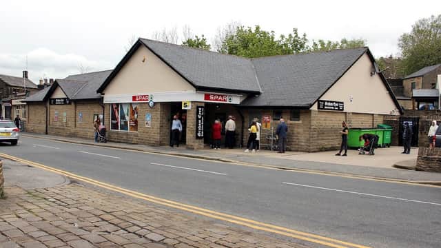 SPAR grocery store in Oxenhope, Yorkshire, with people outside, stone facade, and a green recycling bin nearby.