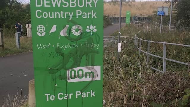 Welcome sign for Dewsbury Country Park with greenery and a path leading to the car park in the background.