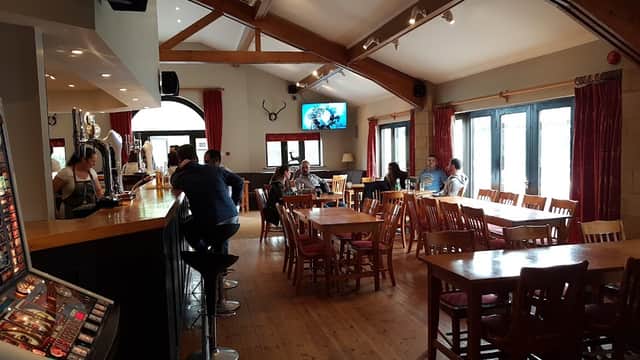 Warm pub interior with wooden beams, tables, and patrons enjoying drinks. A TV displays a sports game in the background.