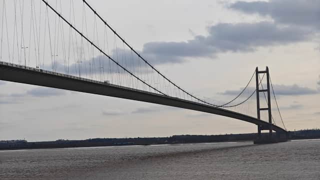 The Humber Bridge stretches across a cloudy sky, towering over the muddy riverbanks below.