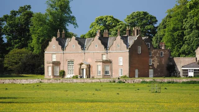 Historic stone house with large windows, surrounded by lush greenery and a vibrant yellow flower field.