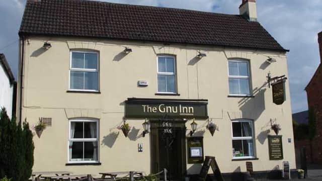 The Gnu Inn pub features a cream-colored exterior, wooden signage, and outdoor seating with flower baskets.