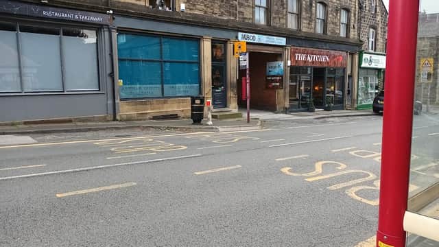 Street view of Cross Roads Park with shops, a bus stop, and parked cars in a quaint Yorkshire setting.
