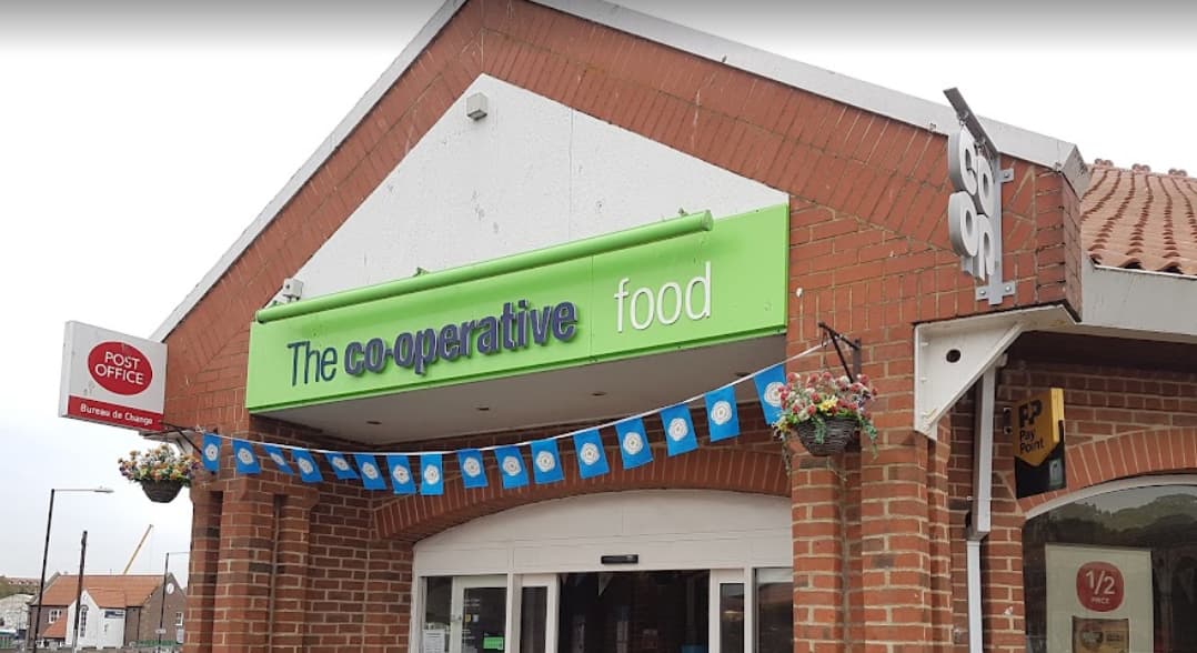 Co-op Food store entrance with green signage, hanging flower baskets, and a Post Office sign nearby.