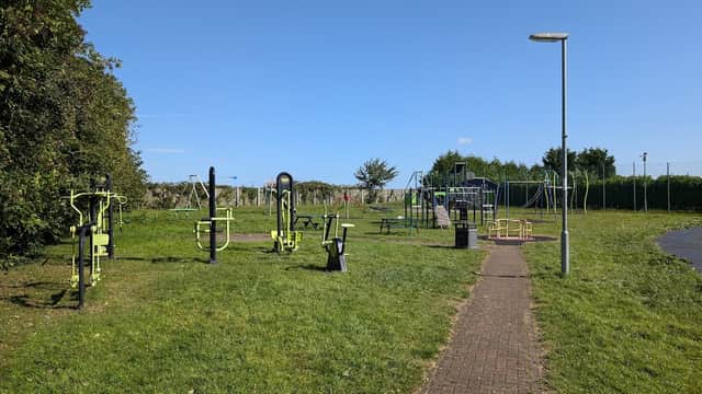 Outdoor play area with exercise equipment, playground structures, benches, and green grass under a clear blue sky.