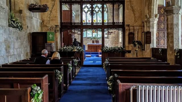 Interior of St Mary's Church with wooden pews, blue carpet, and stained glass windows. Floral decorations line the aisle.