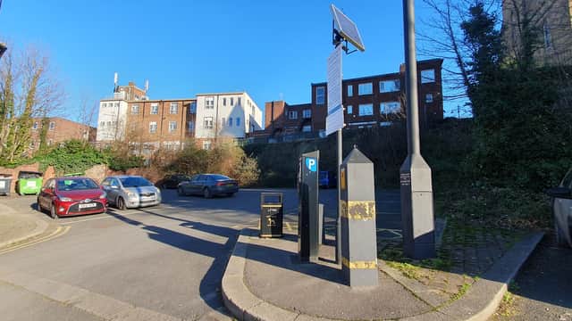 Pay & Display car park with parked cars, a payment machine, and buildings in the background under a clear blue sky.